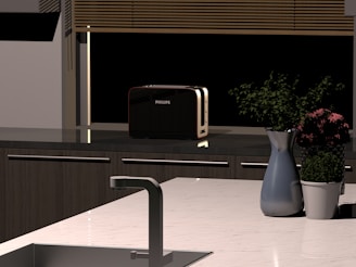 A modern kitchen countertop featuring a metal sink, a silver faucet, and a sleek black toaster. A vase with purple flowers and a green plant in a white pot are also placed on the counter. Blinds cover a window in the background.