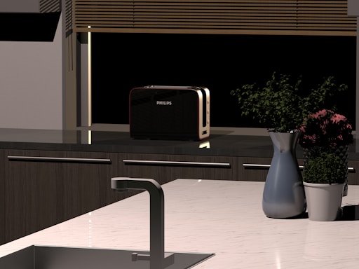 A modern kitchen countertop featuring a metal sink, a silver faucet, and a sleek black toaster. A vase with purple flowers and a green plant in a white pot are also placed on the counter. Blinds cover a window in the background.