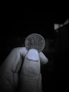 A close-up view of a person's hand holding an Indian coin between their thumb and index finger. The coin features the Lion Capital of Ashoka engraved on it, with the inscriptions 'भारत' and 'INDIA' on either side. The background is dark and out of focus, highlighting the coin and the hand holding it.