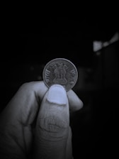 A close-up view of a person's hand holding an Indian coin between their thumb and index finger. The coin features the Lion Capital of Ashoka engraved on it, with the inscriptions 'भारत' and 'INDIA' on either side. The background is dark and out of focus, highlighting the coin and the hand holding it.