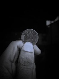 A close-up view of a person's hand holding an Indian coin between their thumb and index finger. The coin features the Lion Capital of Ashoka engraved on it, with the inscriptions 'भारत' and 'INDIA' on either side. The background is dark and out of focus, highlighting the coin and the hand holding it.