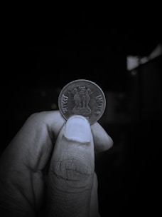 A close-up view of a person's hand holding an Indian coin between their thumb and index finger. The coin features the Lion Capital of Ashoka engraved on it, with the inscriptions 'भारत' and 'INDIA' on either side. The background is dark and out of focus, highlighting the coin and the hand holding it.