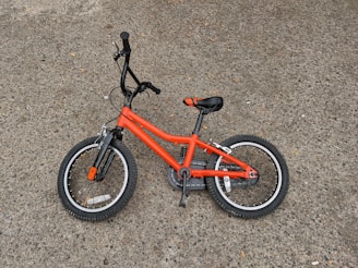 A small orange child's bicycle lying on a rough gray pavement. The bike has black handlebars, pedals, and seat, with orange accents on the tires.