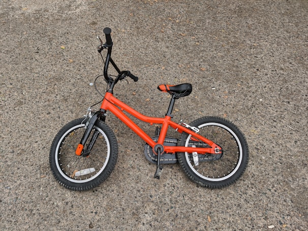 A small orange child's bicycle lying on a rough gray pavement. The bike has black handlebars, pedals, and seat, with orange accents on the tires.