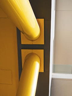 Two large yellow pipes aligned horizontally against a structural wall, with clear geometric shapes and sharp angles in the background.
