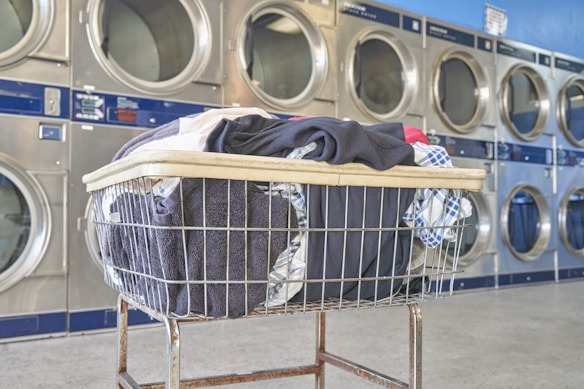 A laundry cart filled with various clothing items sits in front of a row of industrial washers in a laundromat. The scene is brightly lit and features several round washer doors reflecting the light.