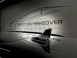 A sleek and glossy surface reflecting a stylized design of a car with the words 'Spot On Makeover Car Care' in a minimalistic and modern font. The image includes a focus on a small shark-fin antenna, emphasizing a clean and sophisticated automotive theme.