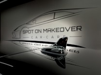 A sleek and glossy surface reflecting a stylized design of a car with the words 'Spot On Makeover Car Care' in a minimalistic and modern font. The image includes a focus on a small shark-fin antenna, emphasizing a clean and sophisticated automotive theme.