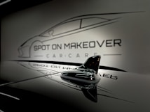 A sleek and glossy surface reflecting a stylized design of a car with the words 'Spot On Makeover Car Care' in a minimalistic and modern font. The image includes a focus on a small shark-fin antenna, emphasizing a clean and sophisticated automotive theme.