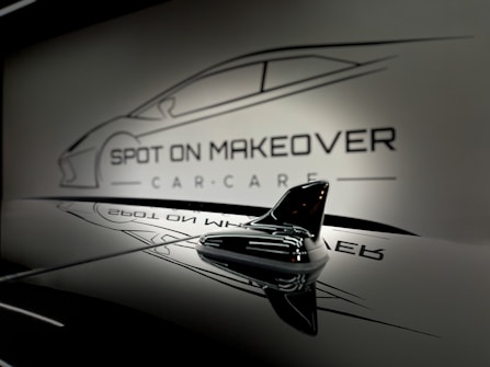 A sleek and glossy surface reflecting a stylized design of a car with the words 'Spot On Makeover Car Care' in a minimalistic and modern font. The image includes a focus on a small shark-fin antenna, emphasizing a clean and sophisticated automotive theme.
