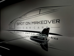 A sleek and glossy surface reflecting a stylized design of a car with the words 'Spot On Makeover Car Care' in a minimalistic and modern font. The image includes a focus on a small shark-fin antenna, emphasizing a clean and sophisticated automotive theme.