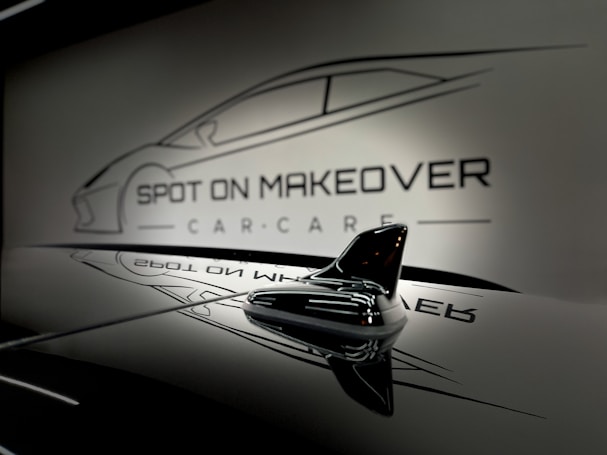 A sleek and glossy surface reflecting a stylized design of a car with the words 'Spot On Makeover Car Care' in a minimalistic and modern font. The image includes a focus on a small shark-fin antenna, emphasizing a clean and sophisticated automotive theme.