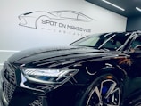 A sleek, modern black car with a shiny finish is parked inside a garage or showroom. The car's detailed and reflective surface highlights the meticulous care given to its maintenance. Above the car, there is a stylized logo and the text 'Spot on Makeover Car Care' on the wall, indicating a specialized vehicle detailing or care service.