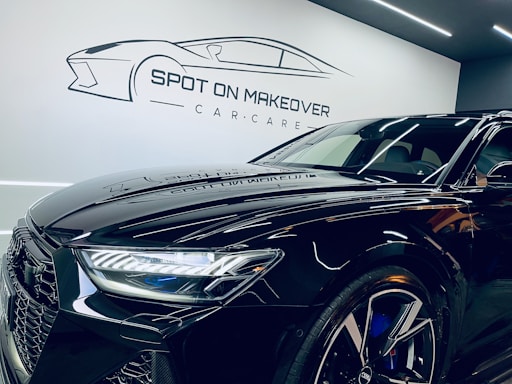 A sleek, modern black car with a shiny finish is parked inside a garage or showroom. The car's detailed and reflective surface highlights the meticulous care given to its maintenance. Above the car, there is a stylized logo and the text 'Spot on Makeover Car Care' on the wall, indicating a specialized vehicle detailing or care service.