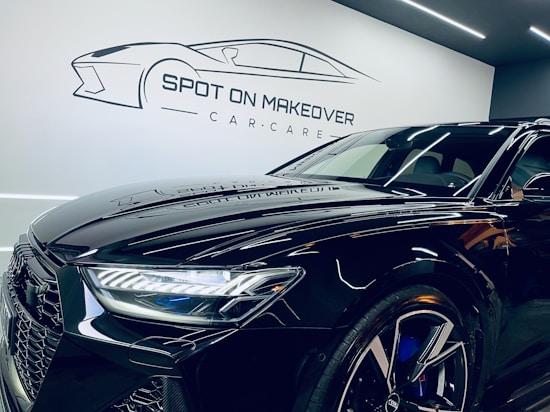 A sleek, modern black car with a shiny finish is parked inside a garage or showroom. The car's detailed and reflective surface highlights the meticulous care given to its maintenance. Above the car, there is a stylized logo and the text 'Spot on Makeover Car Care' on the wall, indicating a specialized vehicle detailing or care service.