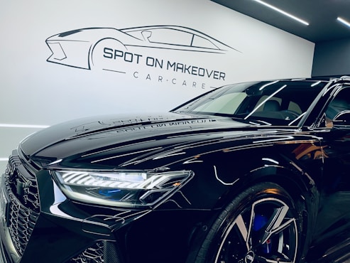 A sleek, modern black car with a shiny finish is parked inside a garage or showroom. The car's detailed and reflective surface highlights the meticulous care given to its maintenance. Above the car, there is a stylized logo and the text 'Spot on Makeover Car Care' on the wall, indicating a specialized vehicle detailing or care service.