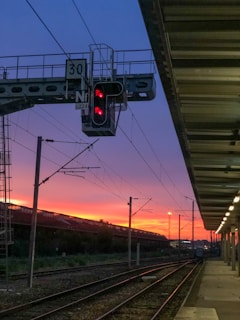 A railway station platform during sunset, with vibrant orange and purple hues in the sky. A railway signal with red lights is prominent in the foreground, alongside overhead wires and tracks extending into the distance. The platform is lit with a series of lamps lining its roof.