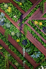 Vertical garden composed of wooden slats arranged in a diagonal, crisscross pattern, with various types of green foliage and vibrant yellow flowers interspersed throughout. Succulents and ferns add texture and diversity to the lush greenery.
