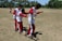 Three children wearing matching red and white outfits are balancing on wooden planks as part of an outdoor team-building activity. They are on a grassy field with trees and a soccer goal in the background.
