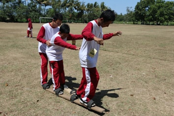 Three children wearing matching red and white outfits are balancing on wooden planks as part of an outdoor team-building activity. They are on a grassy field with trees and a soccer goal in the background.