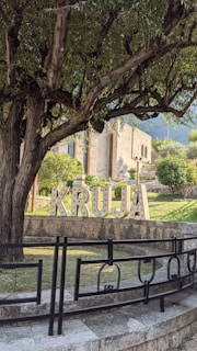 A large tree with dense foliage partially shades a sign spelling 'KRUJA' in decorative letters. The sign is positioned in a lush garden area near ornamental stonework and railings. In the background, a stone building with arched windows is visible, surrounded by more greenery and distant mountains.