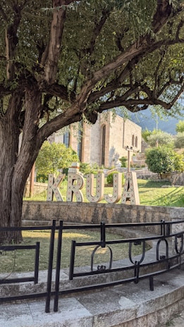 A large tree with dense foliage partially shades a sign spelling 'KRUJA' in decorative letters. The sign is positioned in a lush garden area near ornamental stonework and railings. In the background, a stone building with arched windows is visible, surrounded by more greenery and distant mountains.