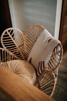 A wicker chair is adorned with an intricate rattan weave design, featuring a cushion in a muted beige tone. Draped over the backrest is a light-colored fabric with a subtle pattern, situated near a wall with soft light casting shadows.