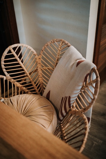 A wicker chair is adorned with an intricate rattan weave design, featuring a cushion in a muted beige tone. Draped over the backrest is a light-colored fabric with a subtle pattern, situated near a wall with soft light casting shadows.