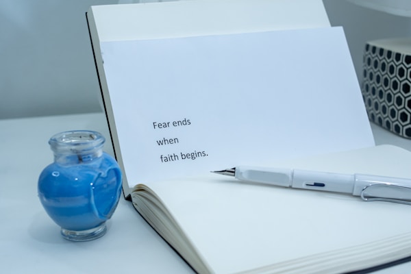 An open notebook displays a page with the text 'Fear ends when faith begins.' Nearby, a pen rests on the pages, and a small glass jar containing blue liquid is placed on the left side. The scene is set on a white tabletop, creating a calm and minimalistic ambiance.
