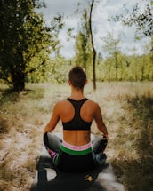 A person with short hair is sitting cross-legged on a yoga mat outdoors in a grassy area, surrounded by trees and sunlight filtering through. They are wearing a black sports bra and dark leggings with a colorful waistband.