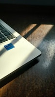 A partial view of a laptop with a visible Intel Core i5 sticker, placed on a dark wooden surface. Sunlight is casting shadows at an angle across both the laptop and the table.