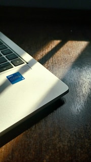A partial view of a laptop with a visible Intel Core i5 sticker, placed on a dark wooden surface. Sunlight is casting shadows at an angle across both the laptop and the table.