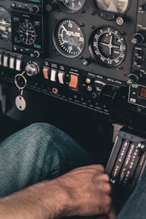 Close-up of an aircraft cockpit showing various gauges, switches, and controls. A hand rests on the throttle quadrant, with jeans visible in the background. The cockpit displays instruments for speed, altitude, and navigation.