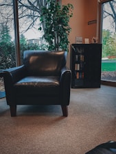 A cozy room featuring a black leather armchair positioned on a carpeted floor with large windows revealing an outdoor view with trees. A leafy green plant stands beside the chair, adding a touch of nature. A small bookshelf filled with books is visible, enhancing the inviting atmosphere.