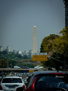 A tall obelisk-like monument is seen in the background, surrounded by urban buildings and foliage. There is a busy multi-lane road with several cars in the foreground. A traffic sign in Portuguese is visible, indicating directions and conditions for drivers.