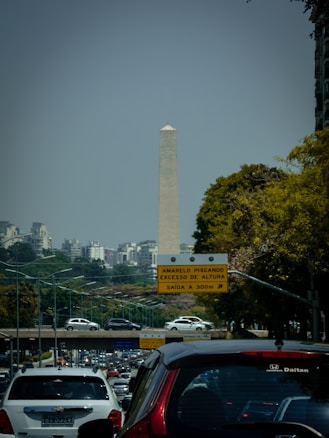 A tall obelisk-like monument is seen in the background, surrounded by urban buildings and foliage. There is a busy multi-lane road with several cars in the foreground. A traffic sign in Portuguese is visible, indicating directions and conditions for drivers.