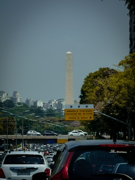 A tall obelisk-like monument is seen in the background, surrounded by urban buildings and foliage. There is a busy multi-lane road with several cars in the foreground. A traffic sign in Portuguese is visible, indicating directions and conditions for drivers.