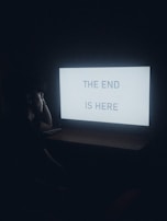 A person sits in a dimly lit room, looking at a glowing computer screen displaying the text 'THE END IS HERE.' The scene is enveloped in shadows, creating a somber and contemplative atmosphere.