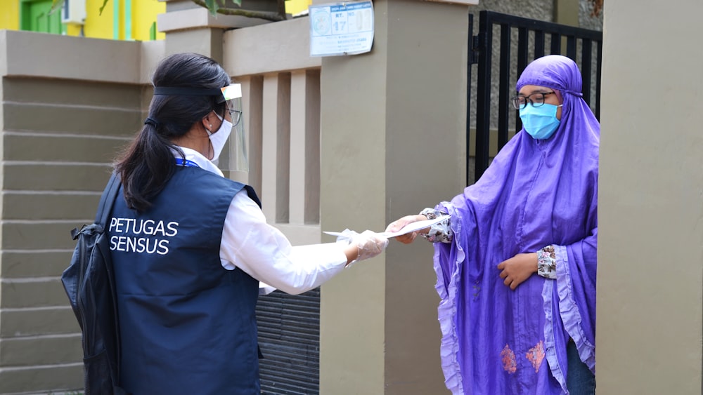 a woman in a purple scarf is handing something to a woman in a blue vest