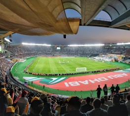 A large, brightly lit stadium filled with spectators. The field is green and well-maintained, with players actively engaged in a soccer match. The stands are crowded, and banners with writing are visible along the perimeter of the field. The atmosphere suggests excitement and energy.