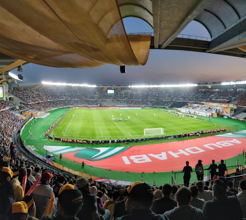 A large, brightly lit stadium filled with spectators. The field is green and well-maintained, with players actively engaged in a soccer match. The stands are crowded, and banners with writing are visible along the perimeter of the field. The atmosphere suggests excitement and energy.
