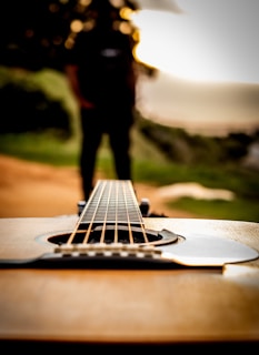 An acoustic guitar lies horizontally, with the focus on its strings and body in the foreground. A blurred figure of a person stands in the background surrounded by natural scenery, suggesting an outdoor setting during sunset or sunrise.