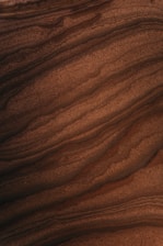 A close-up view of layered rock formations, with smooth, undulating lines creating a textured pattern. The surface is rich in earthy tones and appears to have a natural, organic structure.