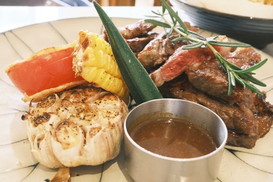 A plate of grilled food including steak with rosemary garnish, roasted garlic, a slice of red bell pepper, a piece of corn on the cob, and a metal container of sauce.
