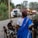 A woman in a blue sari is herding goats along a rural road. She holds a stick in her hand and is surrounded by goats of various colors. In the background, a white SUV is parked near a small village with brick houses and trees.