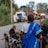 A woman in a blue sari is herding goats along a rural road. She holds a stick in her hand and is surrounded by goats of various colors. In the background, a white SUV is parked near a small village with brick houses and trees.