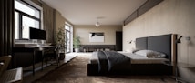 A modern bedroom with minimalist design, featuring a large bed with dark bedding and a sleek headboard. To the left, there's a desk with a computer monitor and a chair. The floor is covered with a textured rug, and a small round side table is positioned next to the bed. The walls are light colored, and a large abstract painting hangs above a low shelf with books. Potted plants are placed near the window and shelf, adding a touch of greenery. Light flows in through large windows with partially drawn curtains.