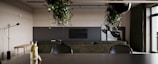 A modern interior space featuring minimalist decor with a dark green sofa facing a mounted flat-screen television. The design includes a mix of natural light and track lighting. Several plants hang from the ceiling, adding a touch of nature. A sleek black floor lamp and a wooden bench are positioned against the wall, and large windows with dark green curtains are on the right.