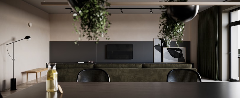 A modern interior space featuring minimalist decor with a dark green sofa facing a mounted flat-screen television. The design includes a mix of natural light and track lighting. Several plants hang from the ceiling, adding a touch of nature. A sleek black floor lamp and a wooden bench are positioned against the wall, and large windows with dark green curtains are on the right.