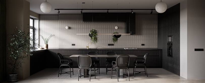 A modern kitchen primarily featuring dark tones with a minimalistic design. There is a large dining table with matching chairs in the center. The kitchen counter is equipped with modern appliances, and potted plants are hanging from a rail as well as placed on the counter, adding a touch of greenery. The ambient lighting includes spherical ceiling lamps, and natural light is streaming in through a window on the left side.
